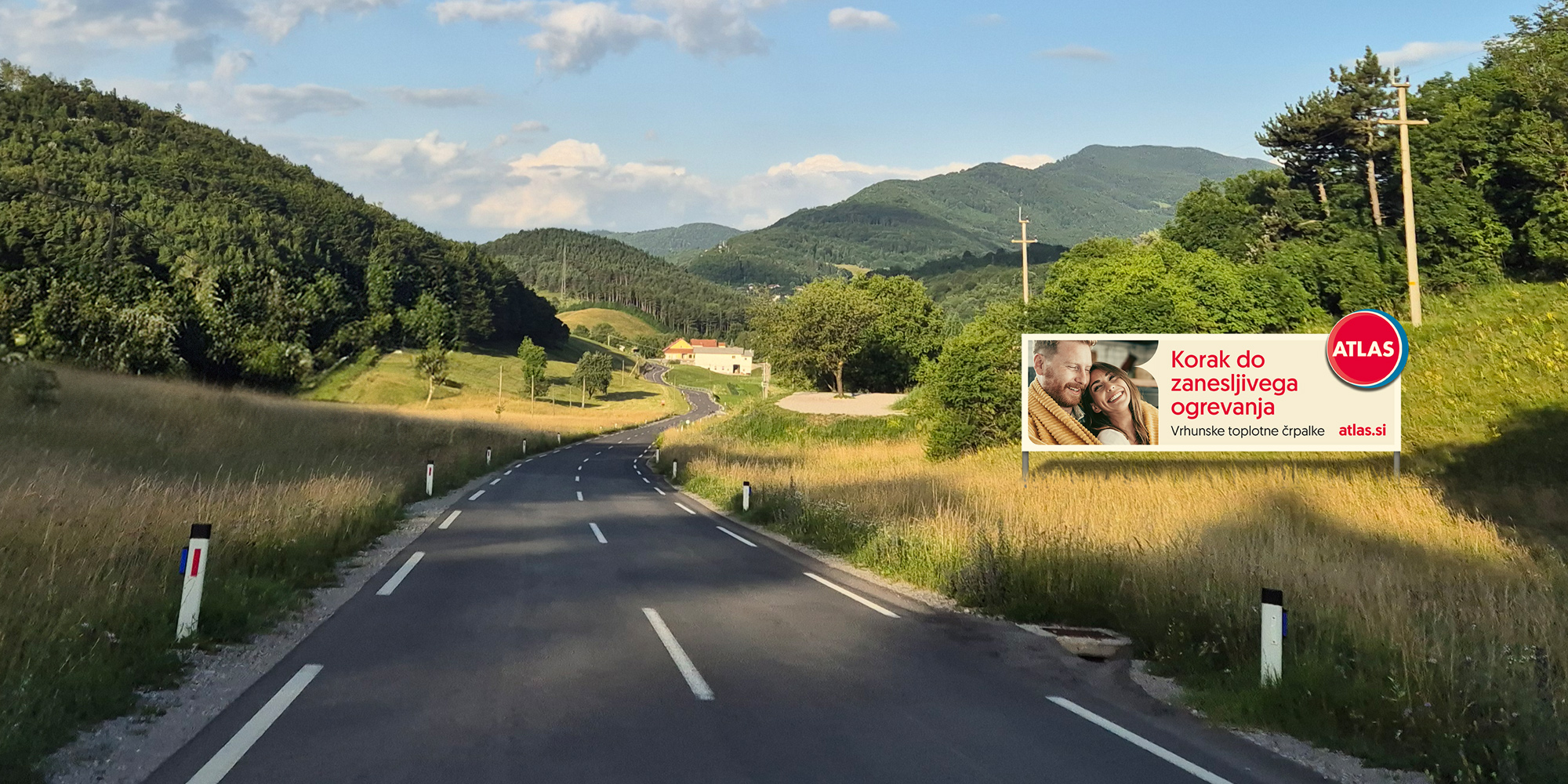 Lokalna cesta, obdana z gozdom in zelenicami na obeh straneh, na levi strani pa velik jumbo plakat z napisom ‘Korak do zanesljivega ogrevanja.