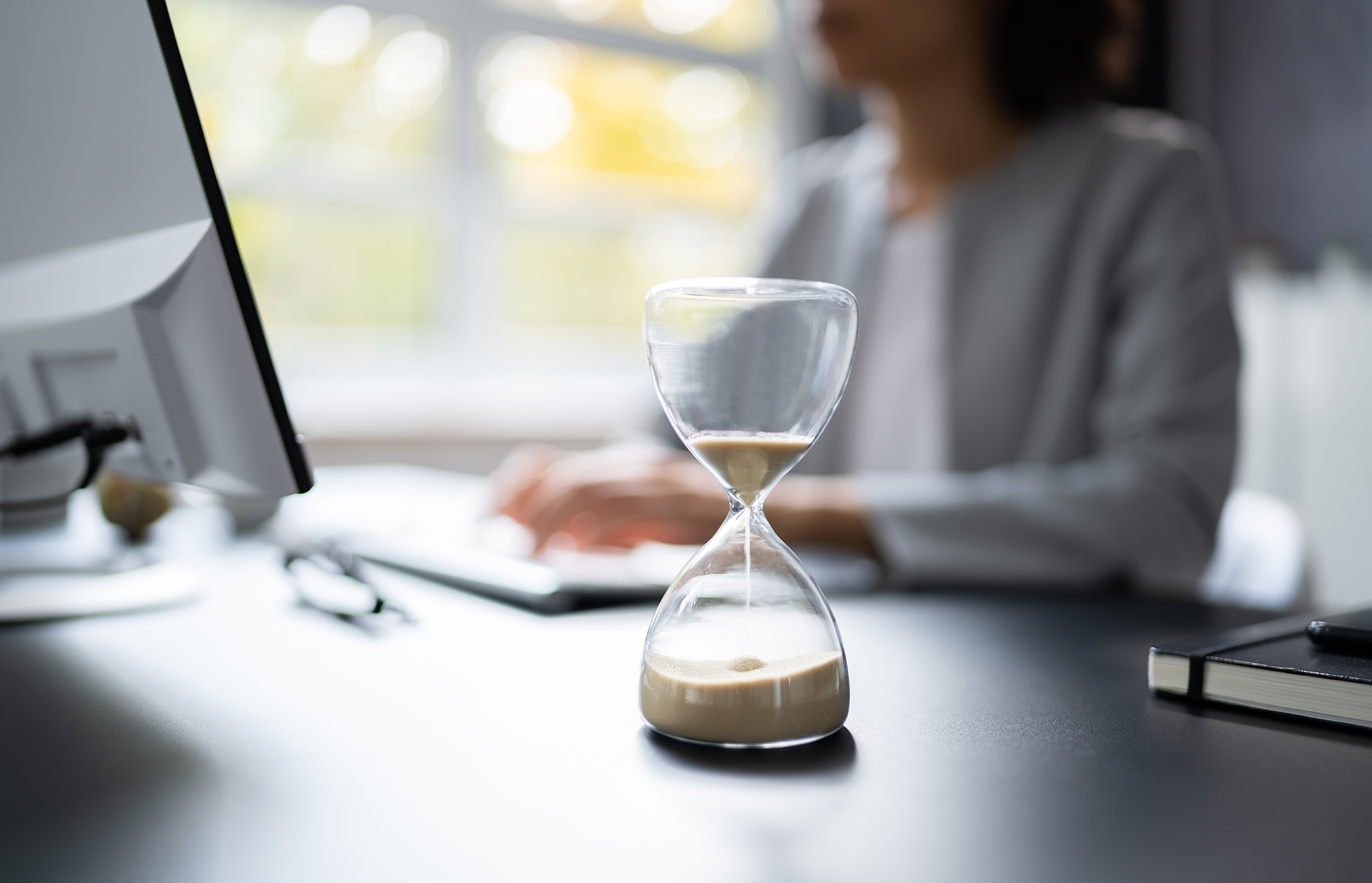 An hourglass with a woman working on a laptop in the background.